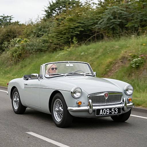 Photograph of a silver classic convertible car with a man in sunglasses driving on a rural road, green foliage in the background. License plate reads 