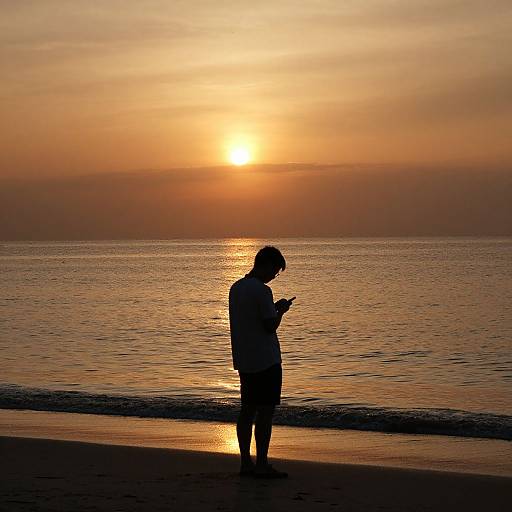 Silhouetted person standing on beach, looking at phone, against vibrant orange sunset over calm ocean. Photograph captures serene, reflective moment.