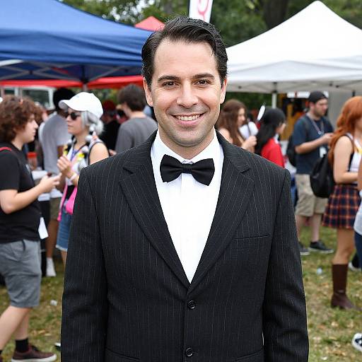 Photograph of a smiling man in a black pinstripe suit with a black bow tie, standing outdoors at a crowded festival with blue and white tents