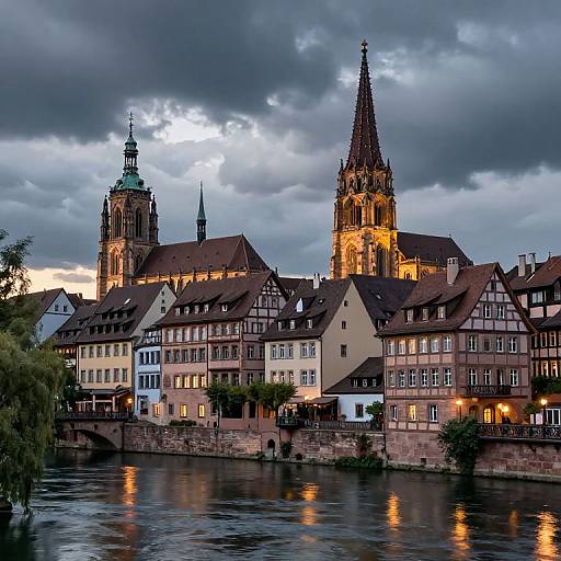 Photograph of a European town at dusk, featuring illuminated Gothic church spires, colorful half-timbered buildings, and a reflective river. Cloud