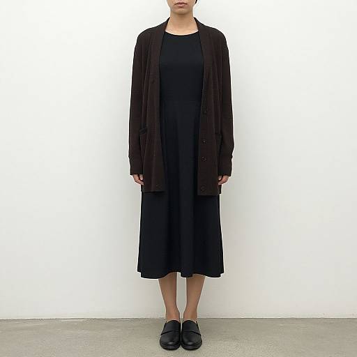 Photograph of a woman in a black dress, dark cardigan, and black loafers, standing against a plain white wall.