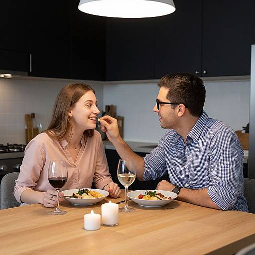 Photograph of a smiling couple dining in a modern kitchen, man feeding woman wine, both with plates, candles, and glasses.