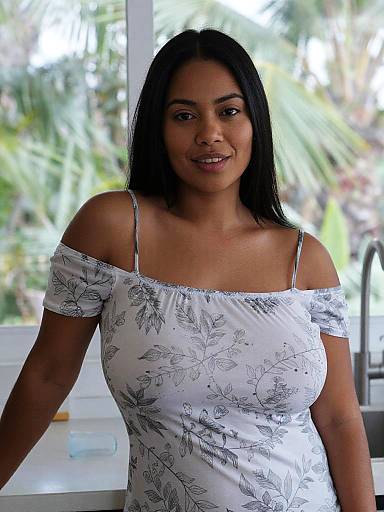 Photograph of a smiling South Asian woman with medium brown skin, black straight hair, wearing a white off-shoulder floral dress, standing indoors with
