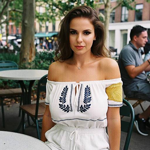Photograph of a young woman with wavy brown hair, wearing an off-shoulder white dress with blue leaf embroidery, seated outdoors at a café