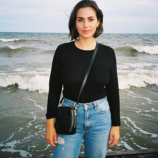 Photograph of a young woman with short black hair, wearing a black long-sleeve top, light blue ripped jeans, and a black crossbody