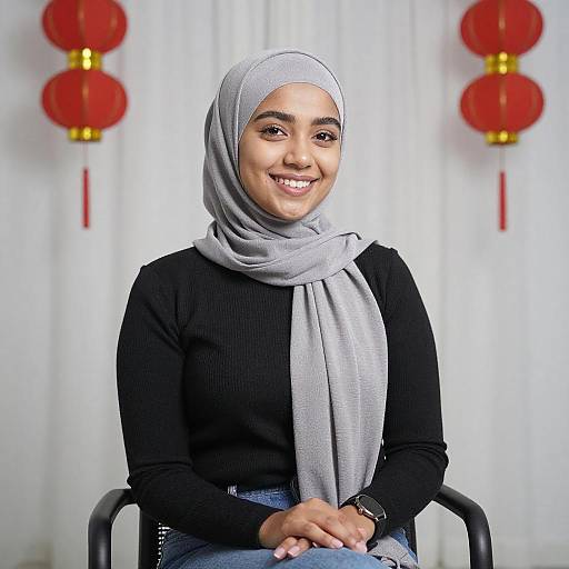 Photograph of a smiling woman with light brown skin, wearing a grey hijab, black long-sleeve top, and grey scarf, seated against