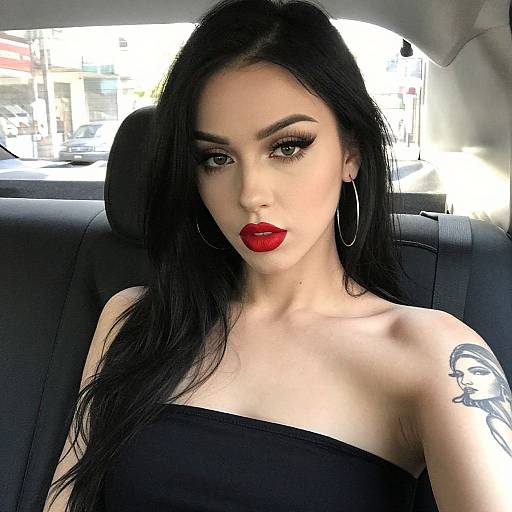 Photograph of a pale-skinned woman with long black hair, red lipstick, hoop earrings, and a tattoo on her right shoulder, sitting in a