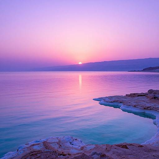 Photograph of a serene sunset over a calm, reflective ocean with pink, purple, and blue hues, and rocky shoreline in the foreground.