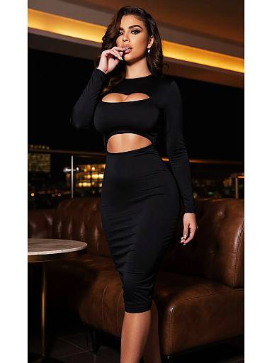 Photograph of a curvy woman with long brown hair, wearing a black cut-out crop top and high-waisted skirt, standing in a dim