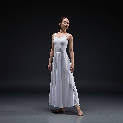 Photograph of a slender woman with fair skin and dark hair in a simple white, sleeveless, flowing dress, standing barefoot against a dark,