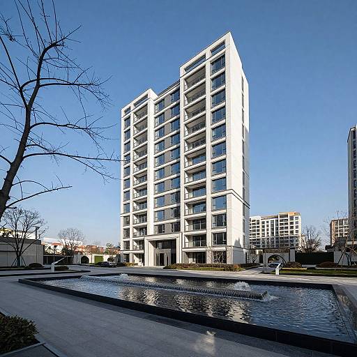 Photograph of a modern, white, 12-story building with large windows, standing beside a reflective water feature on a clear blue day. Bare tree