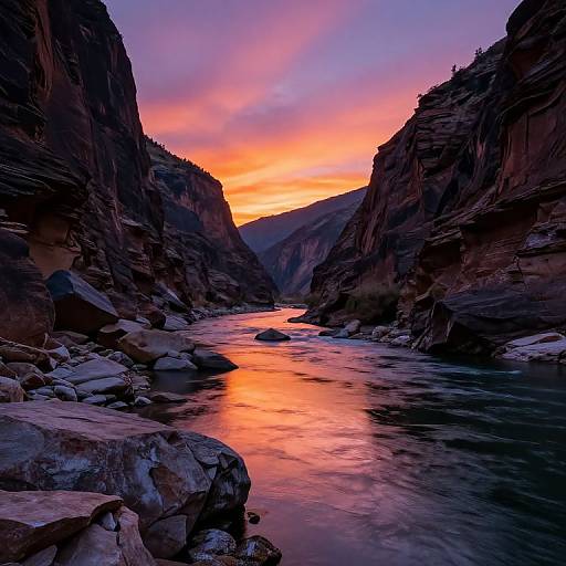 Photograph of a narrow, rocky canyon at sunset with vibrant orange and pink sky, reflecting on a flowing river with smooth stones in the foreground.