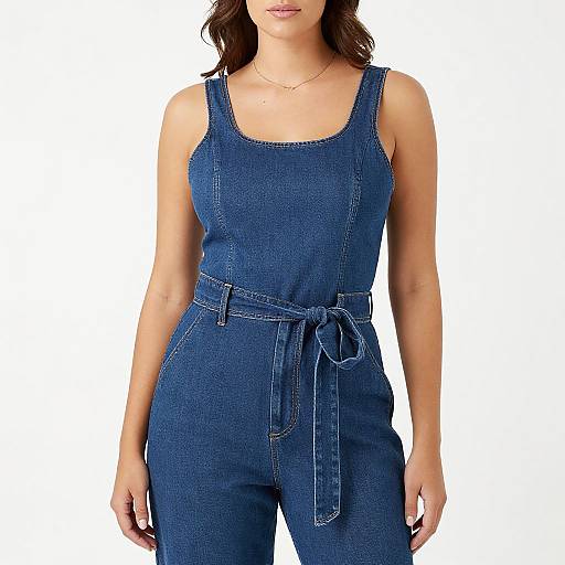 Photograph of a woman with wavy brown hair, wearing a blue denim jumpsuit with a tied waist, white background.