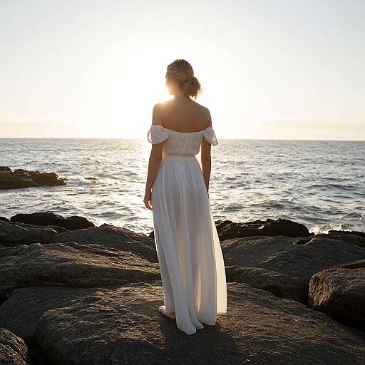 Photograph of a woman in an off-shoulder white dress standing on rocky shoreline, facing the sunlit ocean at sunset.