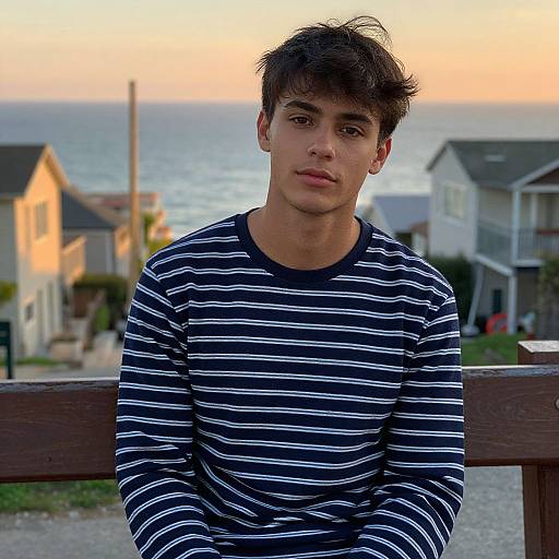 Photograph of a young man with short, dark hair, wearing a black and white striped long-sleeve shirt, leaning on a wooden railing at