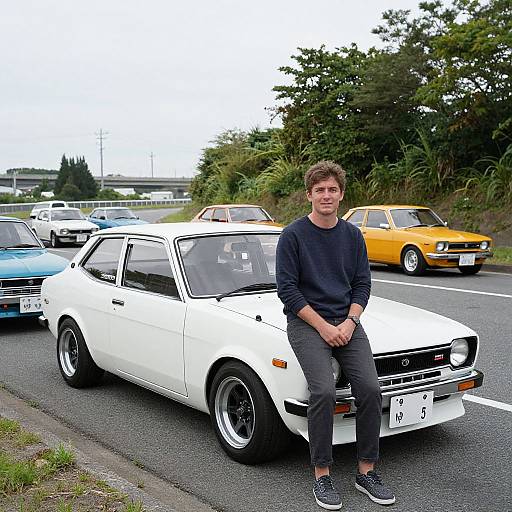 Photograph of a young man with short brown hair, wearing a black sweater and dark pants, sitting on the hood of a white classic car in a