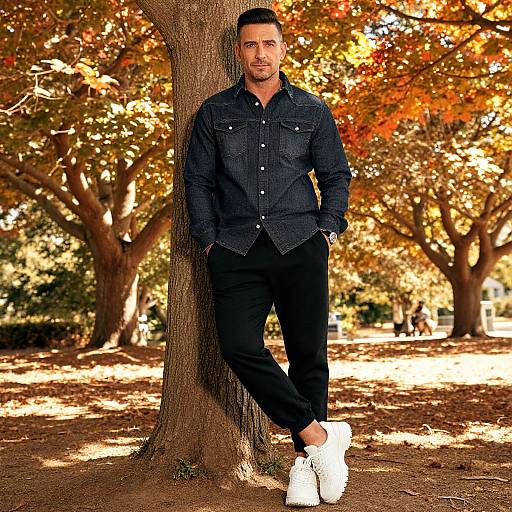 Photograph of a handsome man with short dark hair, wearing a dark denim shirt and black pants, leaning against a tree in a sunlit autumn park