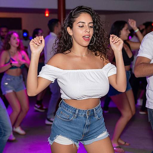 Photograph of a curvy woman with curly black hair, wearing a white off-shoulder crop top and denim shorts, dancing energetically in