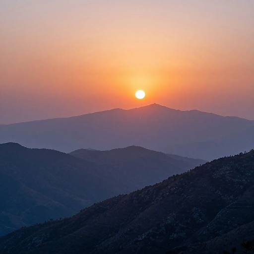 Photograph of a sunset over layered, silhouetted mountain ranges with a glowing orange sun in a gradient sky transitioning from orange to pink to blue