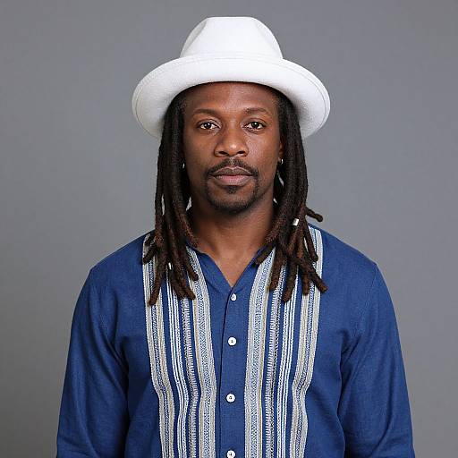 Photograph of a Black man with dreadlocks, wearing a white cowboy hat, blue shirt with white stripes, and neutral gray background.