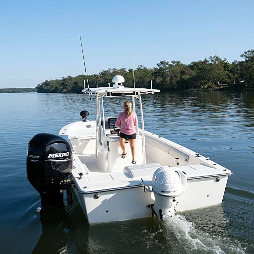 Photograph of a white fishing boat with a woman in pink shirt and black shorts standing at the helm, motor visible, calm water, tree-lined shore