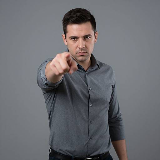 Photograph of a serious-looking, dark-haired man in a gray button-up shirt, pointing directly at the camera against a plain gray background.
