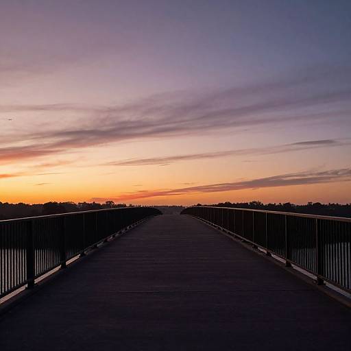 Photograph of a wooden bridge at sunset, with silhouetted railings and a vibrant orange-purple sky, leading into a distant treeline.