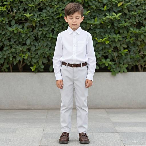 Young boy with short brown hair, wearing a white shirt, pants, brown belt, and brown shoes, standing in front of a green leafy wall