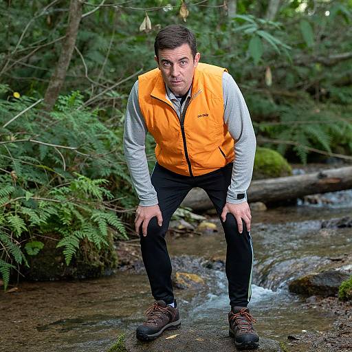 Photograph of a man with short dark hair, wearing an orange vest, grey shirt, black pants, and brown boots, crouching by a