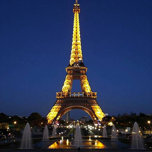 Photograph of the illuminated Eiffel Tower at night, glowing yellow against a deep blue sky, with water fountains in the foreground.