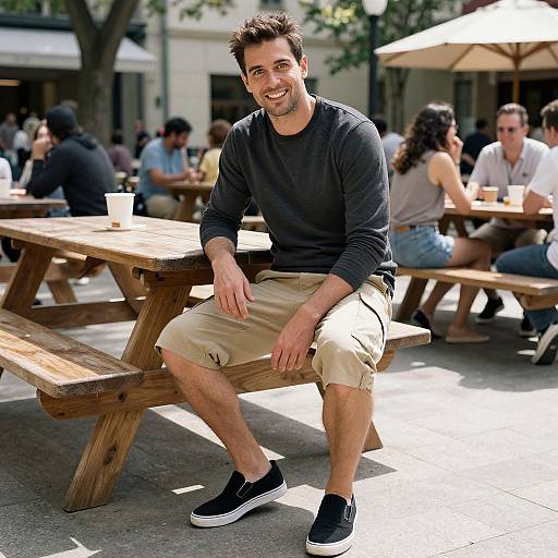 Photograph of a smiling man with short brown hair, wearing a dark long-sleeve shirt, beige knee-length shorts, and black sneakers, sitting