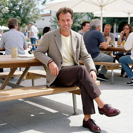 Photograph of a smiling middle-aged man with short brown hair, wearing a checkered blazer, white shirt, and brown pants, sitting on a