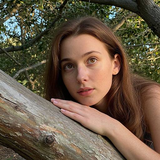 Photograph of a young woman with fair skin and brown hair, resting her head on a weathered tree branch in a sunlit forest. She gaz