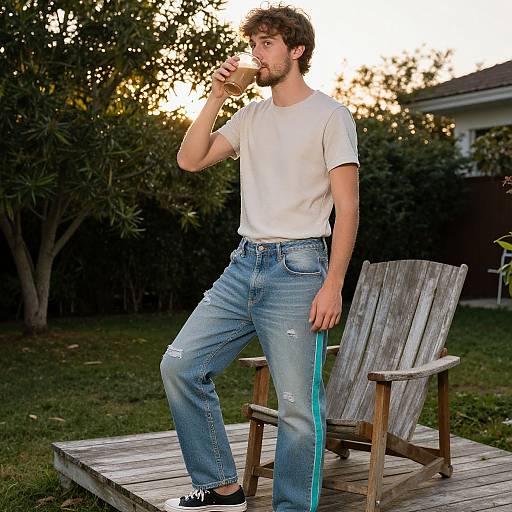 Photograph of a bearded man with curly hair, white t-shirt, ripped blue jeans, black shoes, drinking from a water bottle, standing on