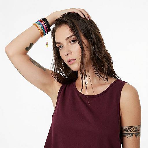 Photograph of a young woman with wet, dark brown hair, wearing a maroon tank top, colorful bracelets, and a tribal tattoo, raising her