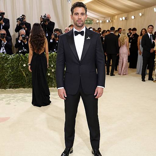 Photograph of a handsome man with short, dark hair and trimmed beard, wearing a black tuxedo and bow tie, standing in a well-l