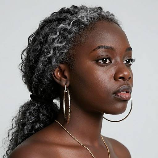 Photograph of a confident, dark-skinned African woman with curly gray-highlighted hair, wearing large gold hoop earrings and a gold necklace, against a
