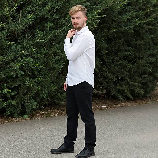 Photograph of a bearded blonde man with short hair, wearing a white shirt, black pants, and black shoes, standing thoughtfully in front of
