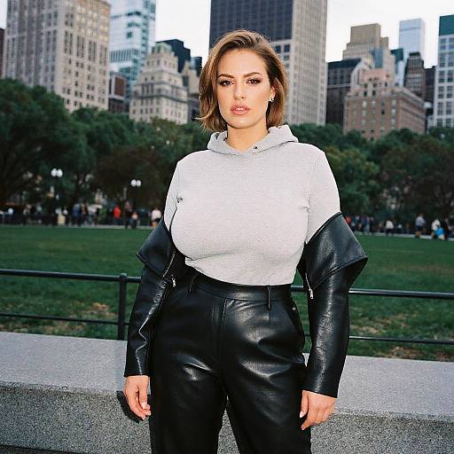 Photograph of a young woman with short brown hair, wearing a white hoodie and black leather pants, standing in a city park with skyscrapers in
