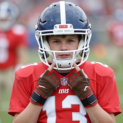 Photograph of a young male football player in a red uniform, black helmet with white stripe, brown gloves, and number 1, looking focused on
