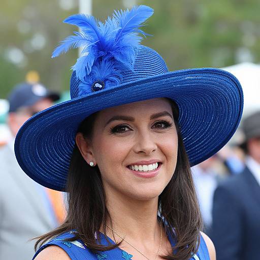 Photograph of a smiling woman with medium brown skin, dark brown hair, wearing a blue feathered hat and matching dress, with blurred background and people