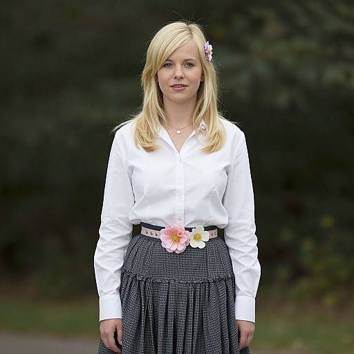 Photograph of a blonde woman with fair skin, wearing a white blouse, black checkered skirt, pink and white flower belt, standing outdoors with a