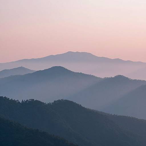 Photograph of misty, layered mountain ranges in varying shades of blue and purple, set against a soft pink and white sky at dawn or dusk.