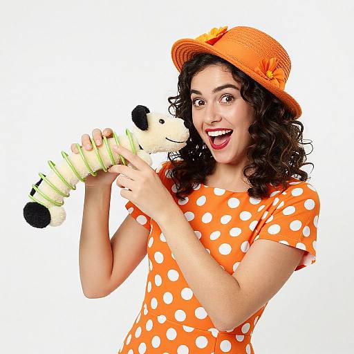 Photograph of a curly-haired woman in an orange polka dot dress, orange hat, smiling, holding a stuffed toy with green accents.