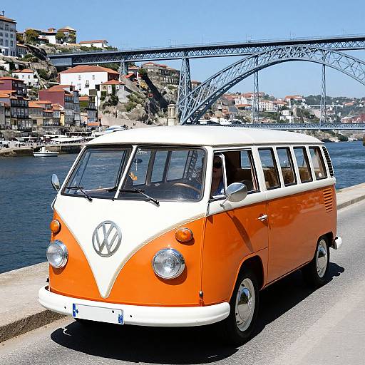 Photograph of an orange and white vintage Volkswagen van parked by a waterfront, with a blue steel bridge and hillside houses in the background under a clear