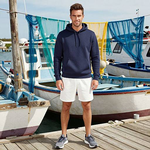 Photograph of a muscular man with short brown hair, wearing a navy hoodie, white shorts, and gray sneakers, standing on a dock with colorful fishing
