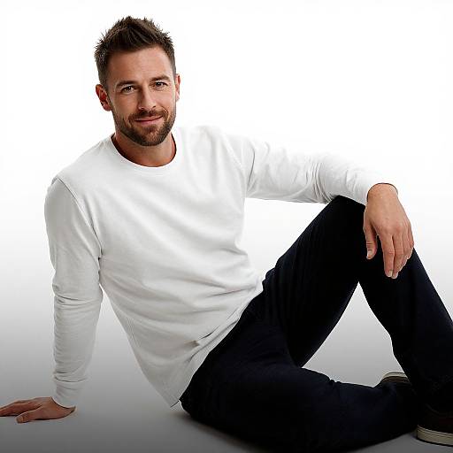 Photograph of a handsome, bearded man with short brown hair, wearing a white long-sleeve shirt and black pants, sitting against a white