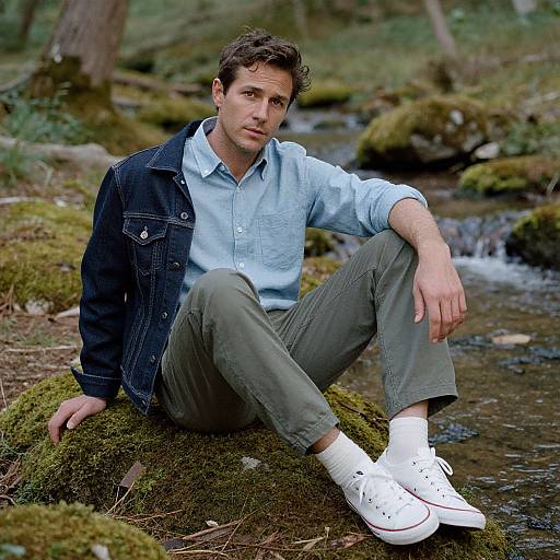Photograph of a young man with curly brown hair, wearing a light blue shirt, dark denim jacket, olive pants, and white sneakers, sitting on