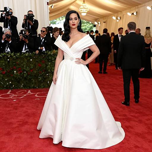 Photograph of a red carpet event featuring a dark-haired woman in a white off-shoulder, V-neck gown, posing confidently with hand on hip