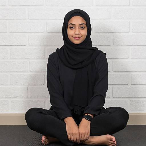 Photograph of a smiling young woman with medium brown skin and dark eyes, wearing a black hijab and outfit, sitting cross-legged on a black mat